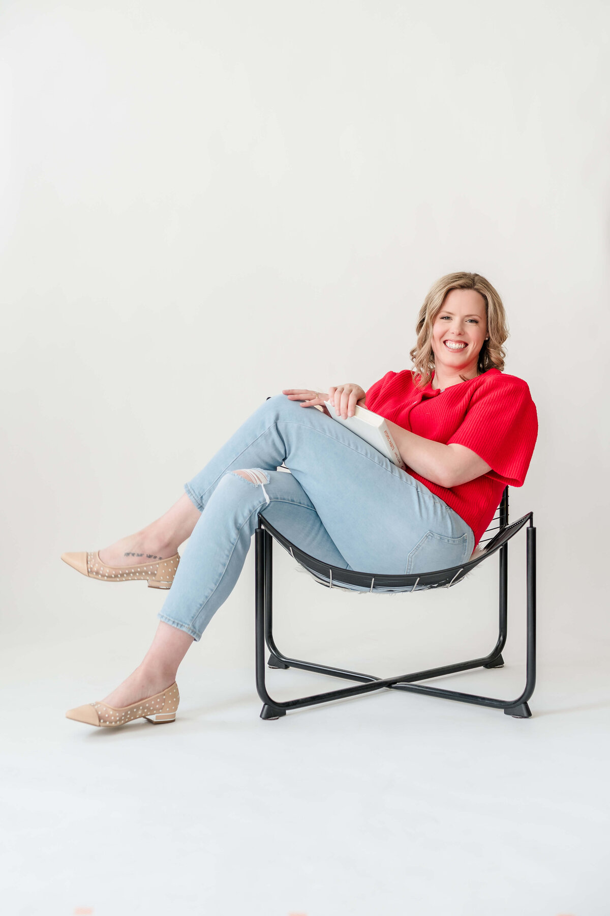 Naturopathic physician lounging in modern chair with book, wearing red knit top and light jeans.