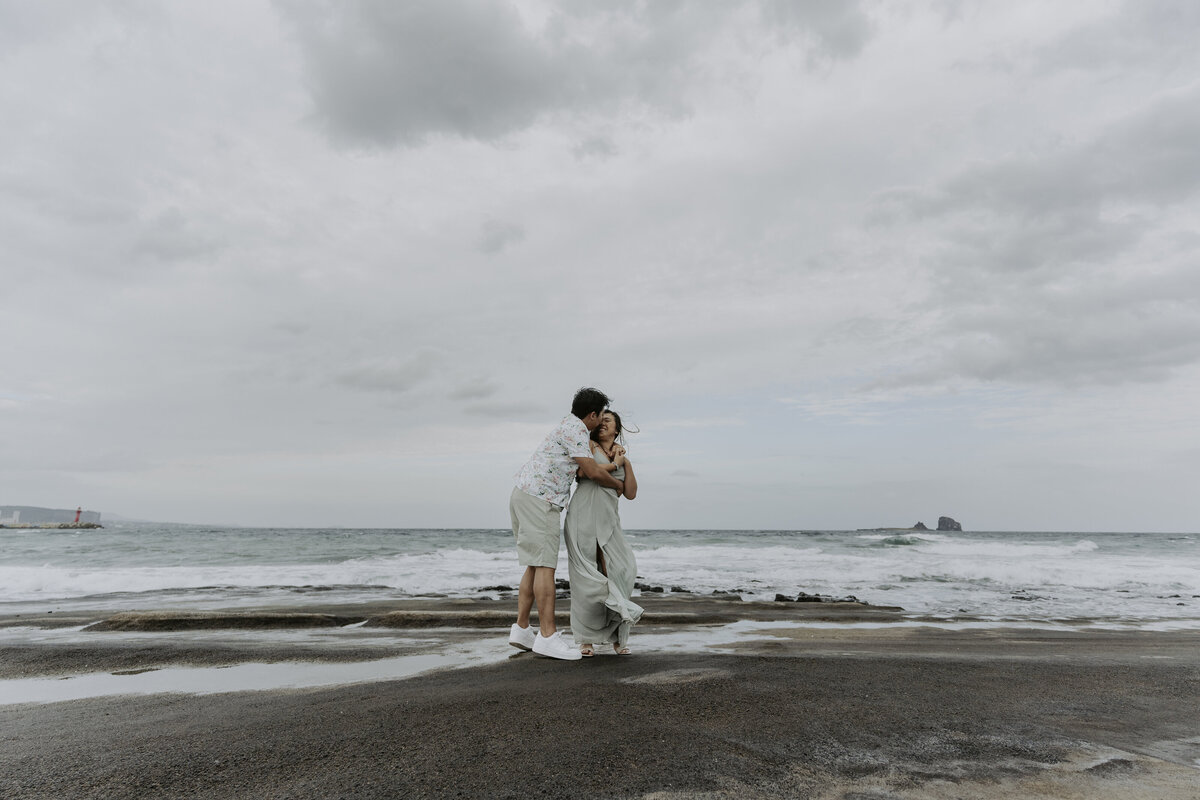 the groom back hugging the bride and kissing her forehead while in the beach of jeju island