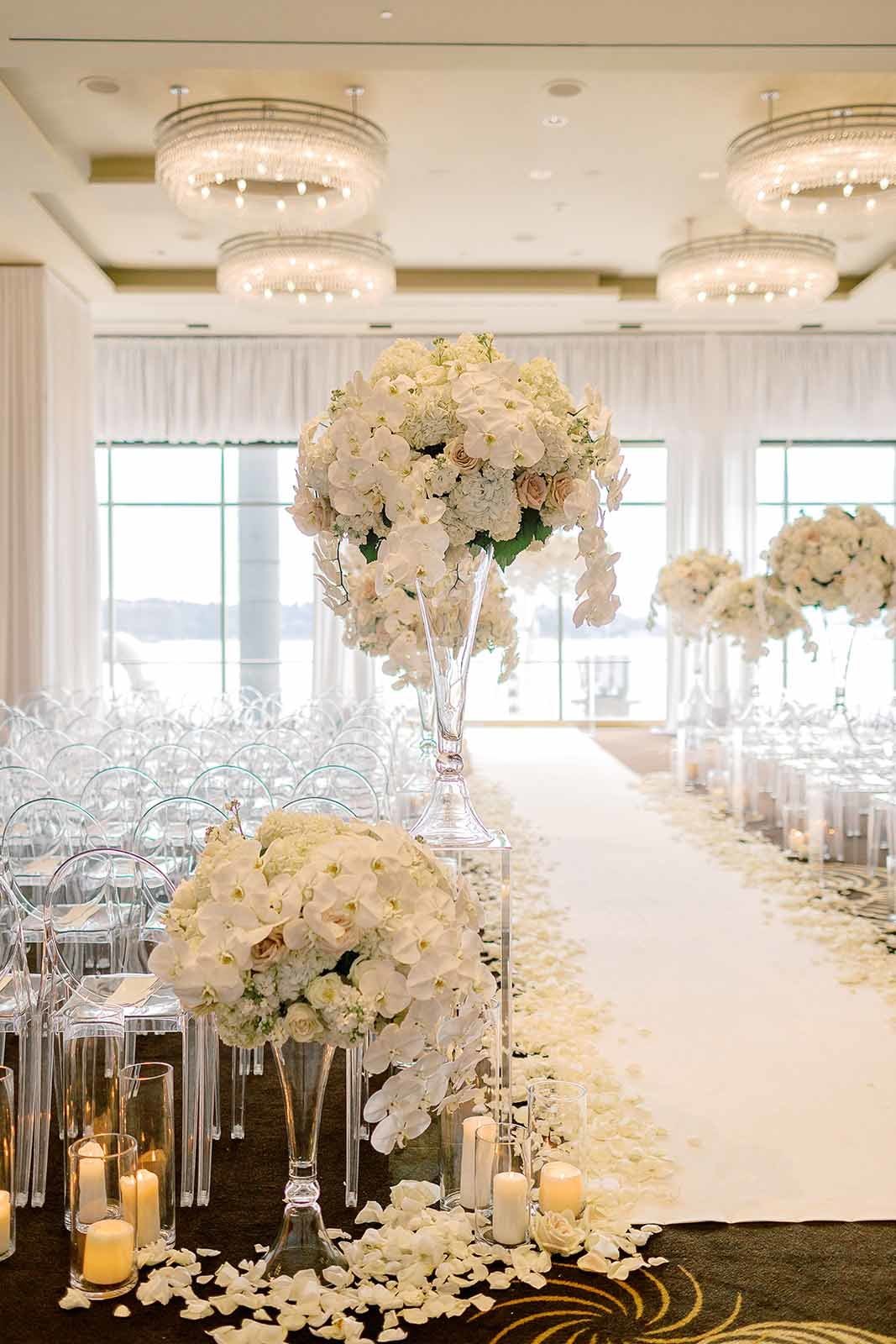 grand wedding ceremony aisle with white flower arrangements on clear lucite pedestals, white rose petals, and hurricane candles lining the aisle