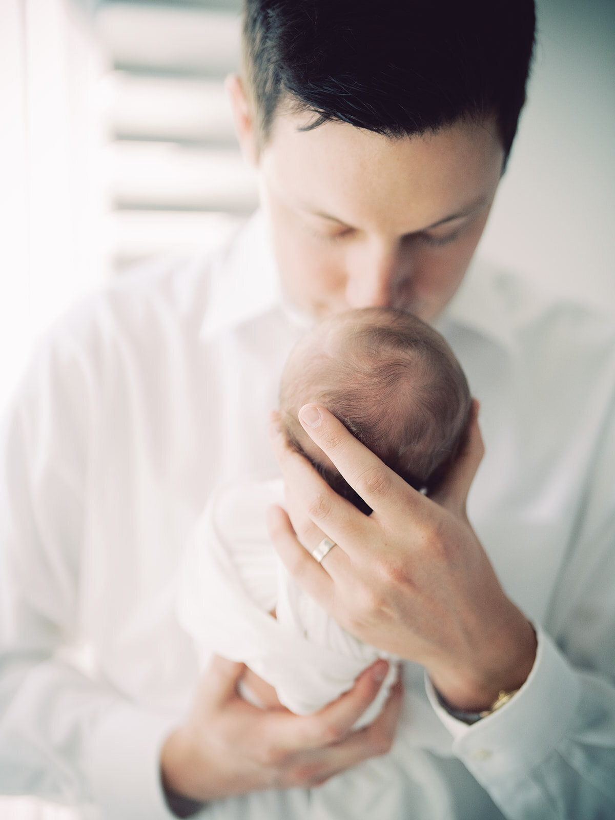 Father in white button-up shirt brings his newborn baby girl up for a kiss.