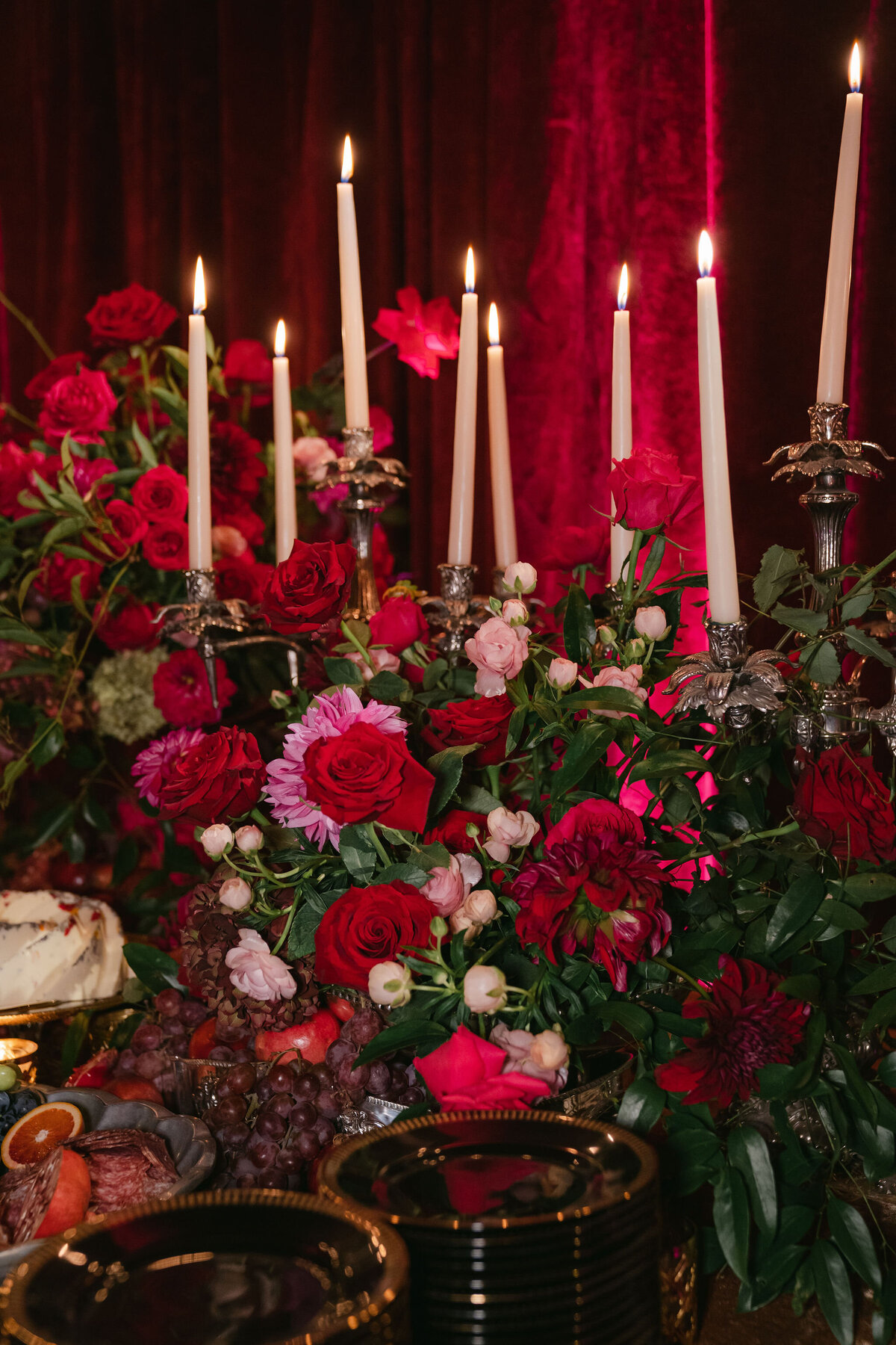 The photo was taken at a tented wedding in Virginia by photographer Elisabetta Marzetti. Clayton Films were the videographers capturing the lush floral arrangement with red, pink, and white blooms illuminated by flickering candles atop an elegant candelabrum. The scene also highlights a thoughtfully decorated table adorned with assorted fruits, a decadent slice of cake, and dark-colored plates, all set against a backdrop of rich red curtains. The overall ambiance exudes an intimate and sophisticated charm.