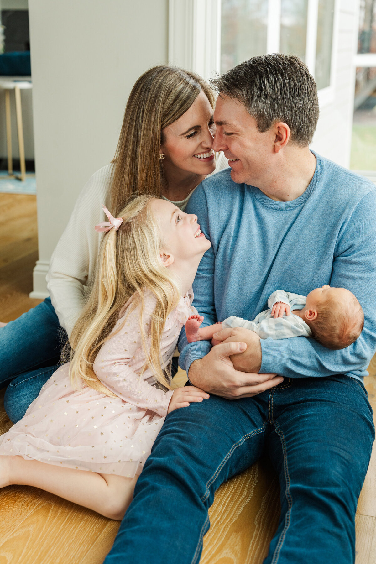 A family sits together on the floor, all gazing warmly at the dad as he cradles the newborn baby