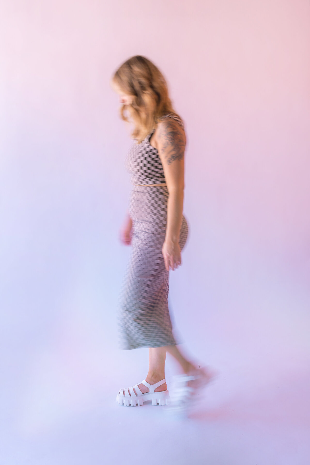 Creative studio branding image featuring a woman in a checkered dress and white platform sandals walking in motion blur across a pink gradient backdrop.