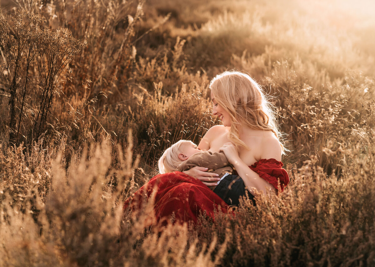Photo of a woman in a red dress sitting in the sunlight breastfeeding her son