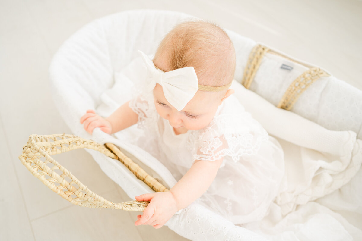 A young baby girl dressed in a white dress with a white bow headband plays inside of a Moses basket during a milestone session in Austin.