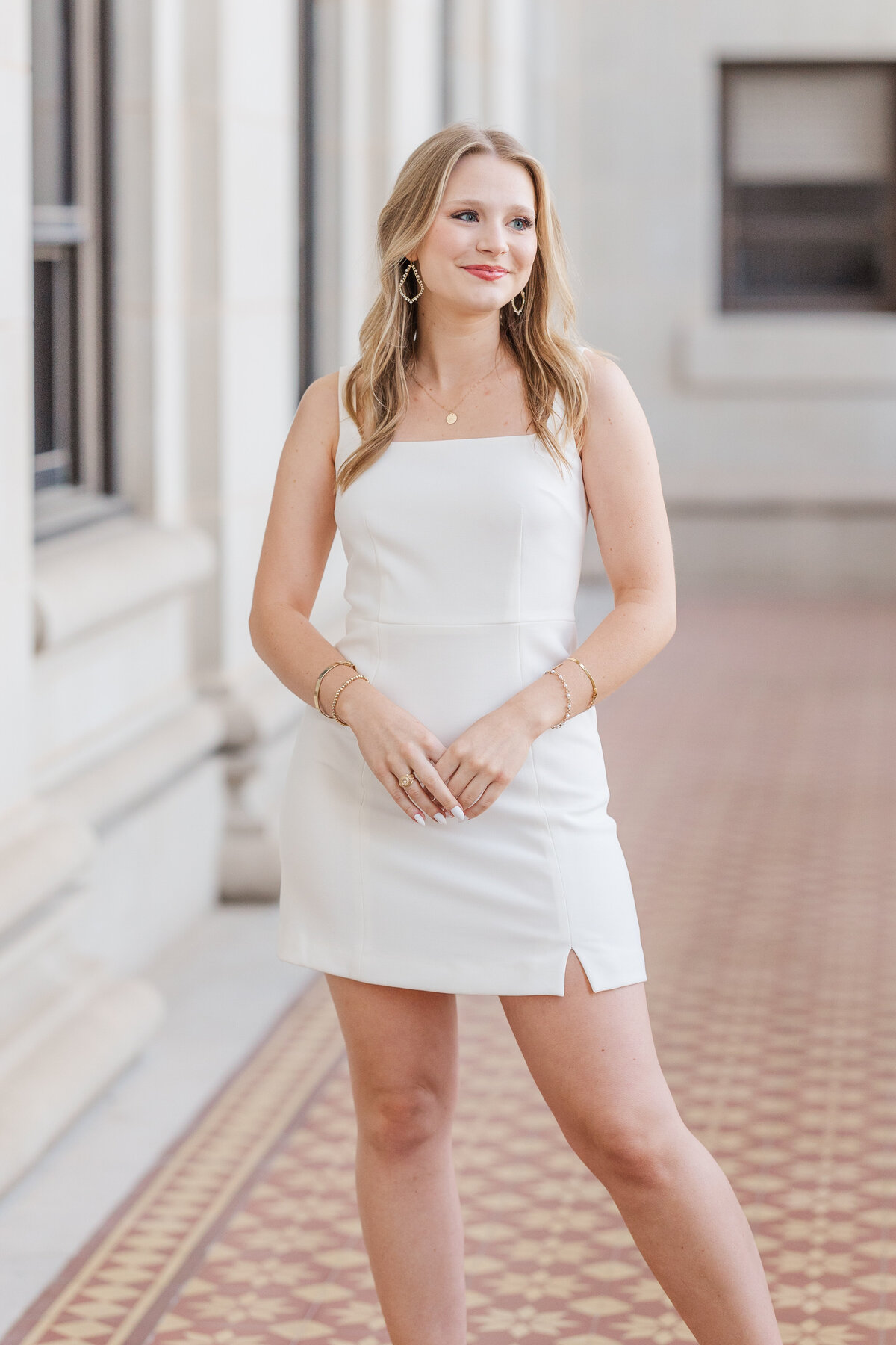Elegant senior portrait of a young woman wearing a white dress, standing in a columned walkway with patterned tile flooring on the Texas A&M University campus in College Station. She smiles confidently, surrounded by timeless architecture.