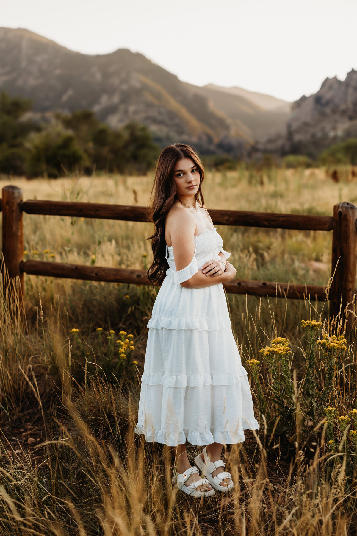 High shool senior girl in white dress standing in front of mountains with tall grasses and yellow flowers