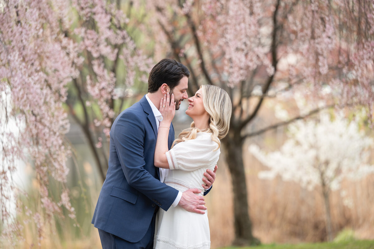 A man and woman stand close together under blooming trees, smiling and touching faces in an outdoor setting.
