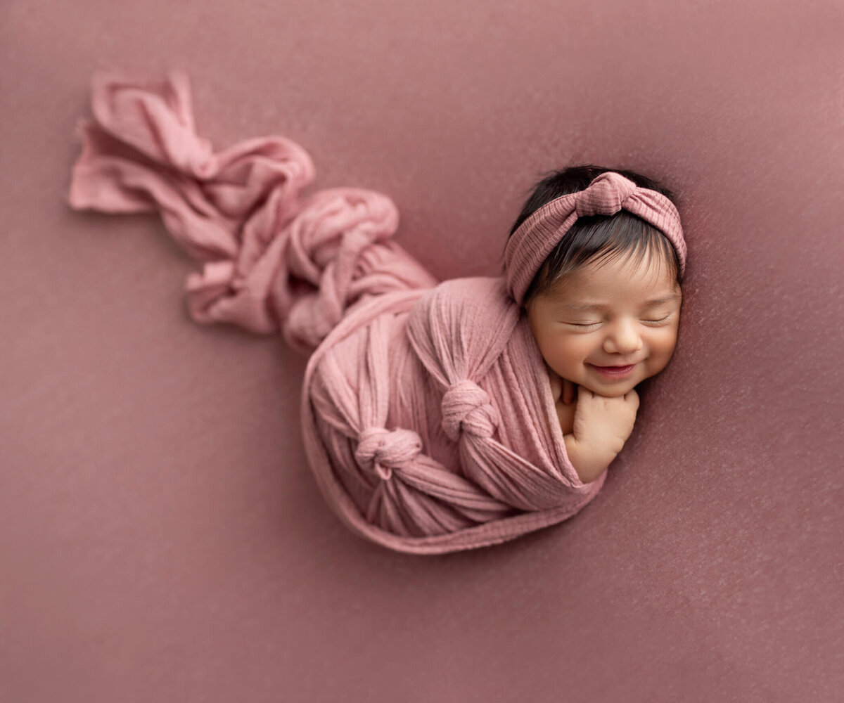 A newborn baby peacefully sleeps, swaddled snugly in a soft pink wrap with knots. The baby wears a matching pink headband and smiles gently in her sleep. The background is a soft pink fabric, creating a warm and serene atmosphere.