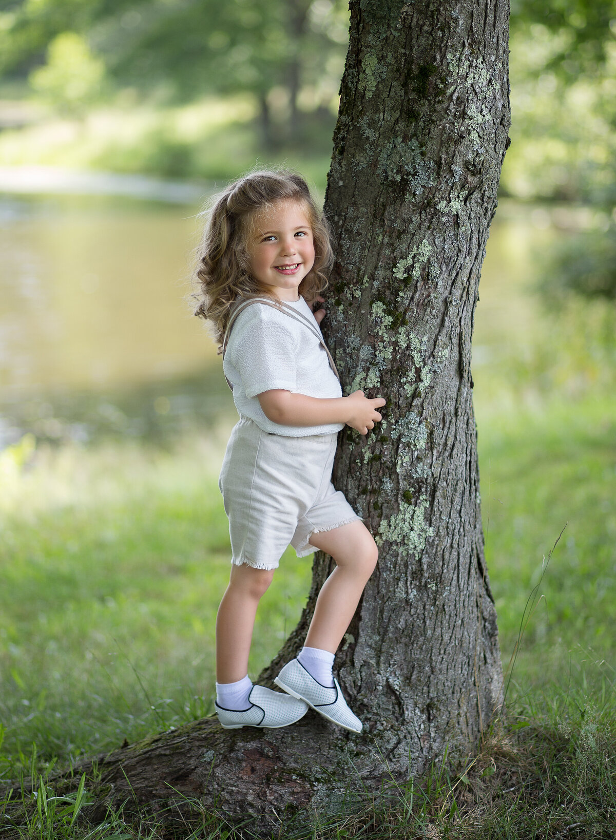 Little boy is standing for a photo to commemorate his first upsherin. He is in a park beside a tree with a big smile facing the camera. Captured by Chaya Bornstein, Brooklyn Family Photographer