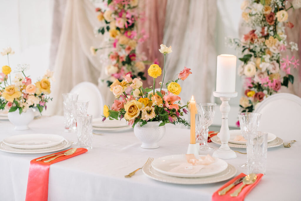 Elegant dining table set with white plates, crystal glasses, and gold cutlery. The centerpiece features vibrant flower arrangements. Two candles, one lit, add to the refined ambiance, perfect for showcasing our wedding planning services that create unforgettable celebrations.