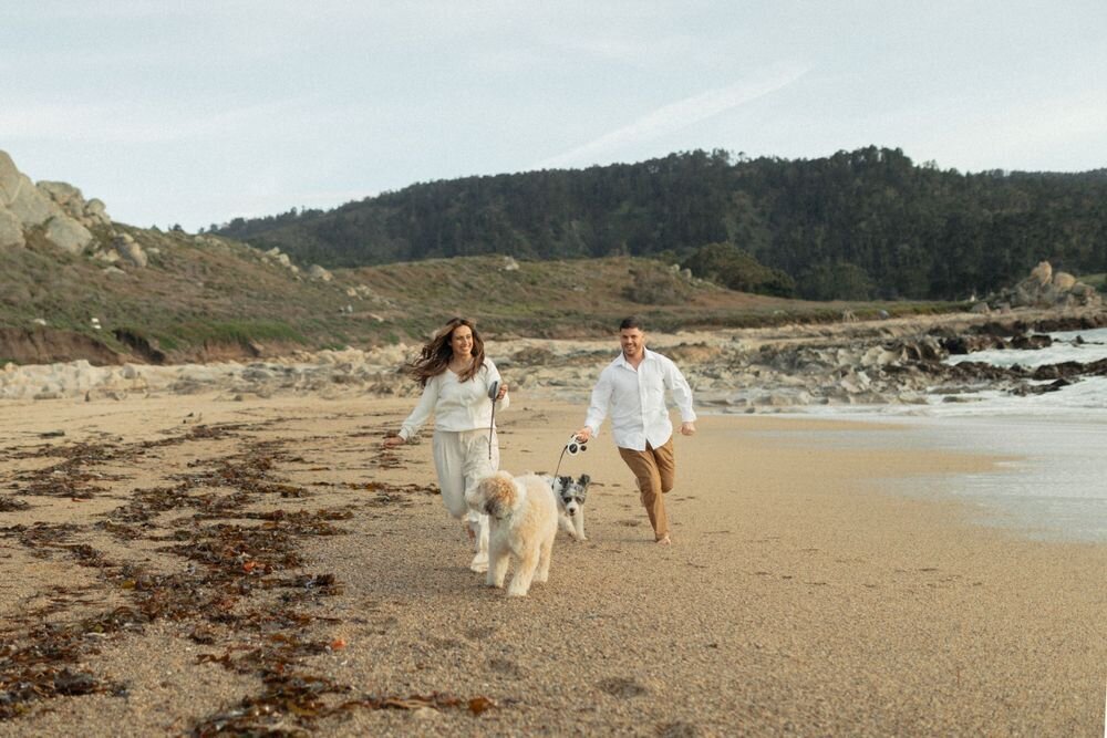 guy and girl running on beach in carmel california on shore with two dogs wearing white outfits during sunset engagement session by katherine krakowski photography