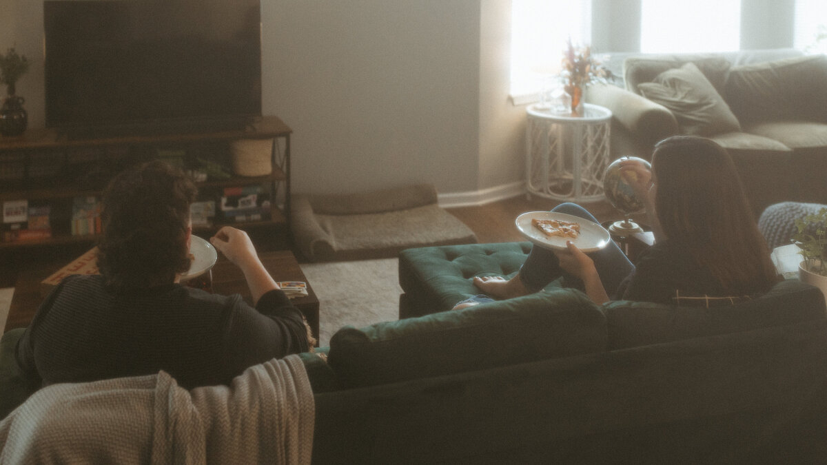 A cozy scene with two people relaxing on a green sofa in a warmly lit living room, one reading a book and the other enjoying a snack