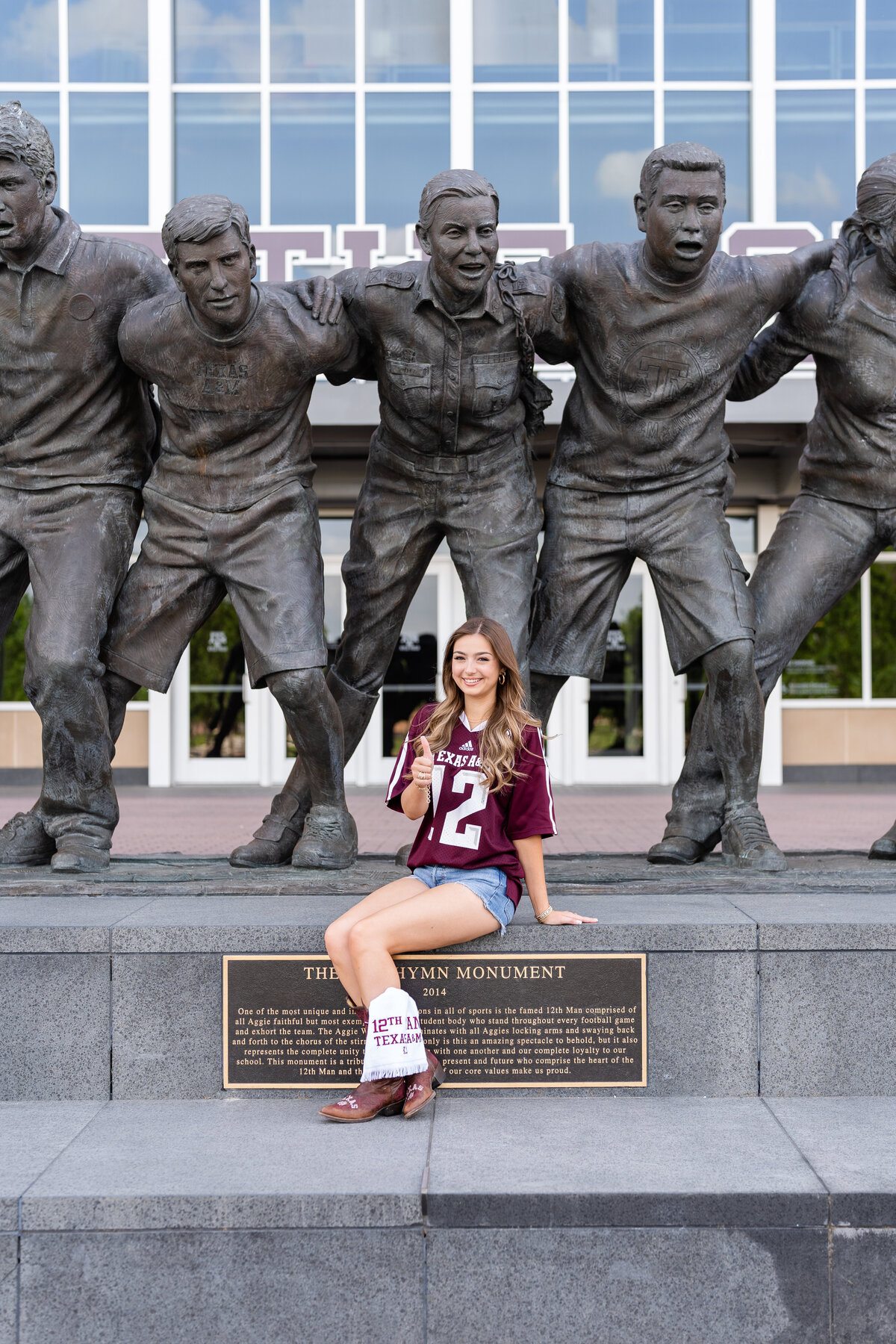 Texas A&M senior girl sitting on war hymn statue and thumbs up wearing a maroon jersey and jean shorts at Kyle Field