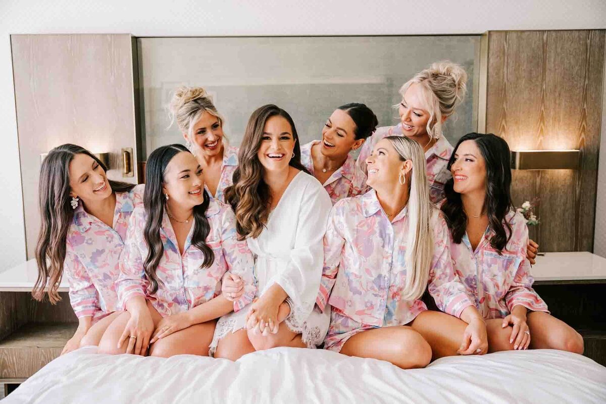 bride is adored by her bridesmaids, while sitting on the bed wearing matching pajamas
