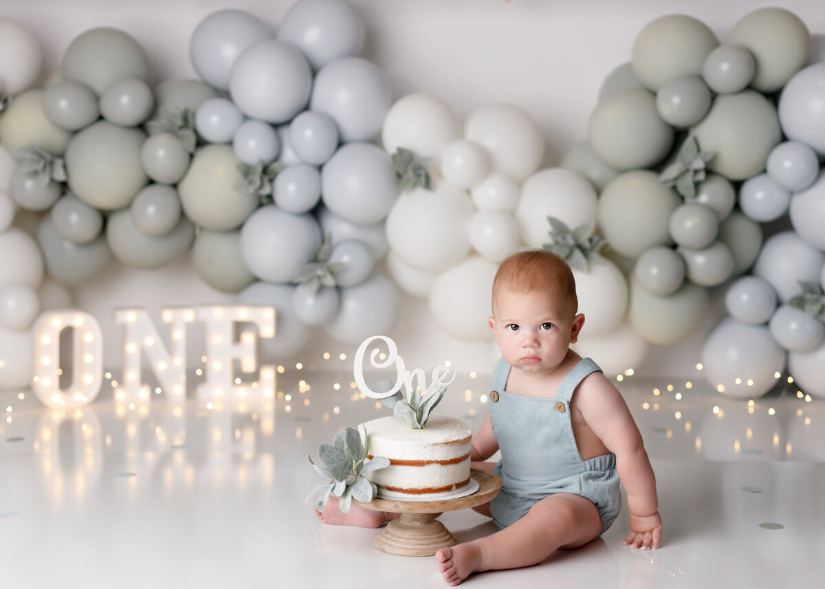 Sage green and light blue balloon garland cake smash in Wellington in West Palm Beach Florida cake smash studio. Baby boy is wearing soft blue overalls sitting beside a white naked cake with greens. The background is Sage, fog blue and cream balloon garland with light up one and lights at the bottom.