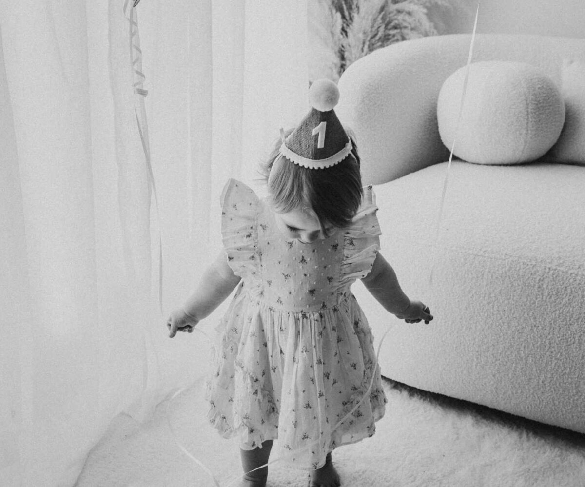 Ruua Hamdan Photography (UK): Black and white image of a baby in a party hat and floral dress looking down.
