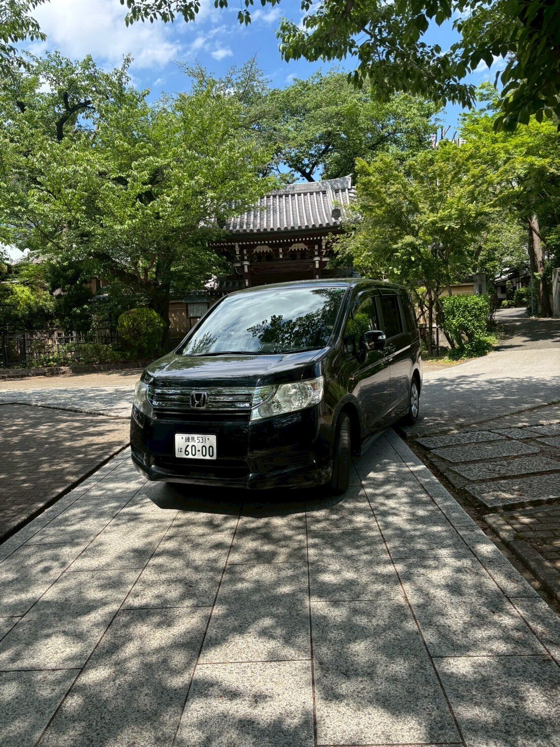A black Honda Stepwgn parked in a serene Japanese temple courtyard surrounded by lush greenery and traditional architecture under a sunny sky.