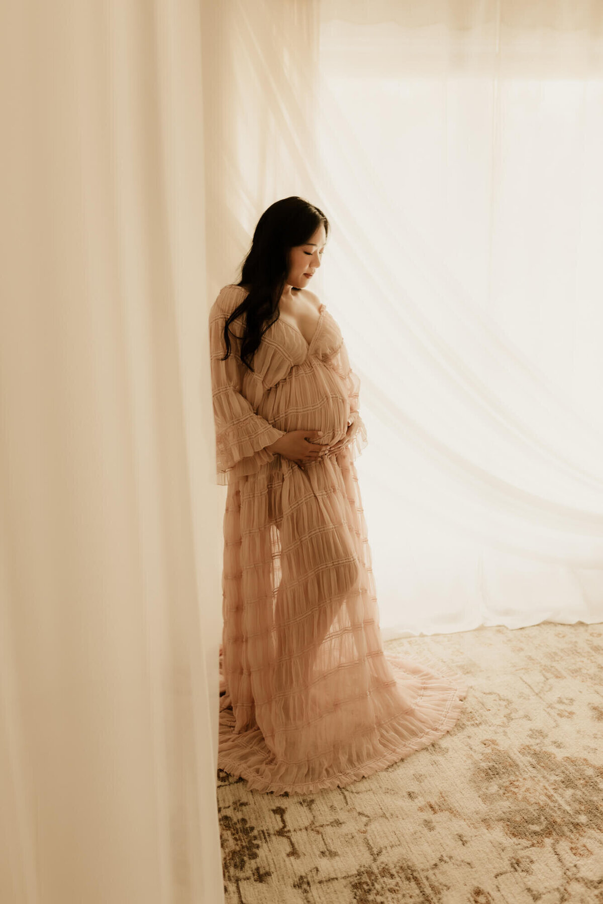 Maternity portrait of a mother looking down at her baby bump while standing and wearing a blush designer dress in Yukon, Oklahoma.