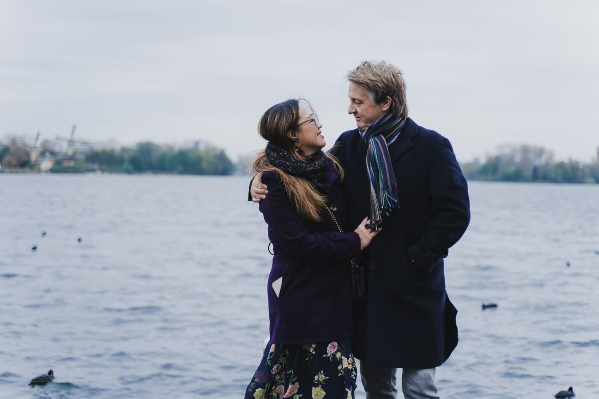 A romantic, full-body portrait of a couple embracing by the water on a cloudy day. The woman, with long brown hair, is wearing glasses, a purple coat, and a floral skirt. She has her arms wrapped around the man, who is wearing a dark blue coat and a striped scarf. They are looking into each other's eyes, and a few ducks are visible on the water behind them.