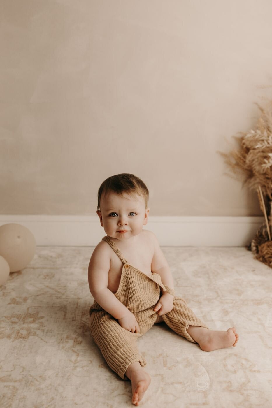 Cute baby sitting on the floor wearing a soft beige color clothes and with a dry plants and balloon at the back