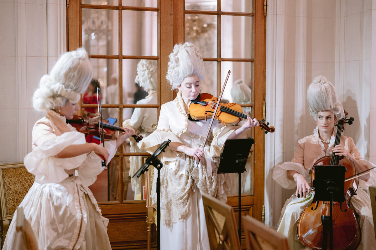 Three musicians dressed in 18th-century attire with elaborate white wigs perform in a refined room. One plays the violin, another also on violin, and the third musician plays the cello. They stand before wooden doors with glass panes. Music stands hold sheet music.