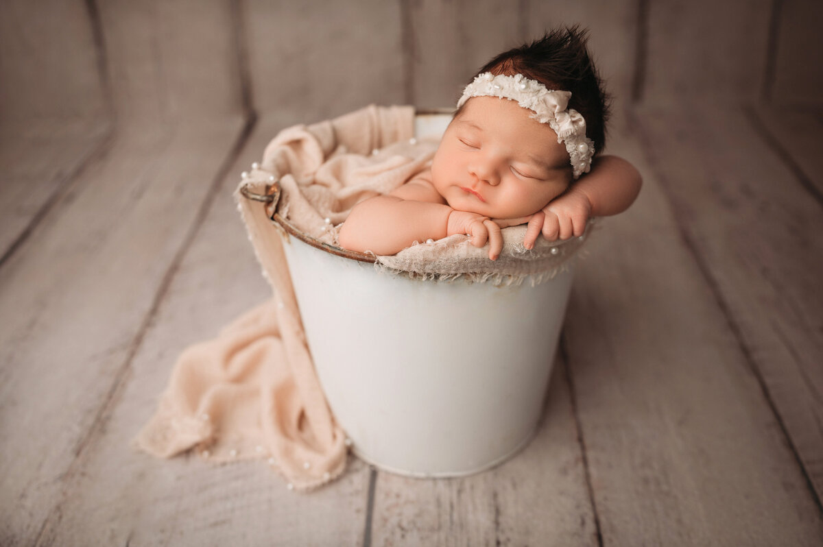 newborn baby girl swaddled in white wrap and wearing a white bow headband posed inside a white heart bowl prop over a white wooden floor