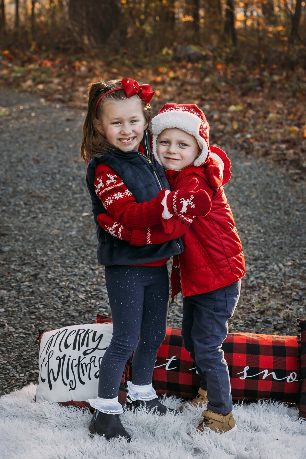brother and sister wearing christmas themed  winter clothing standing on a white fur blanket with xmas pillows and fall leaves