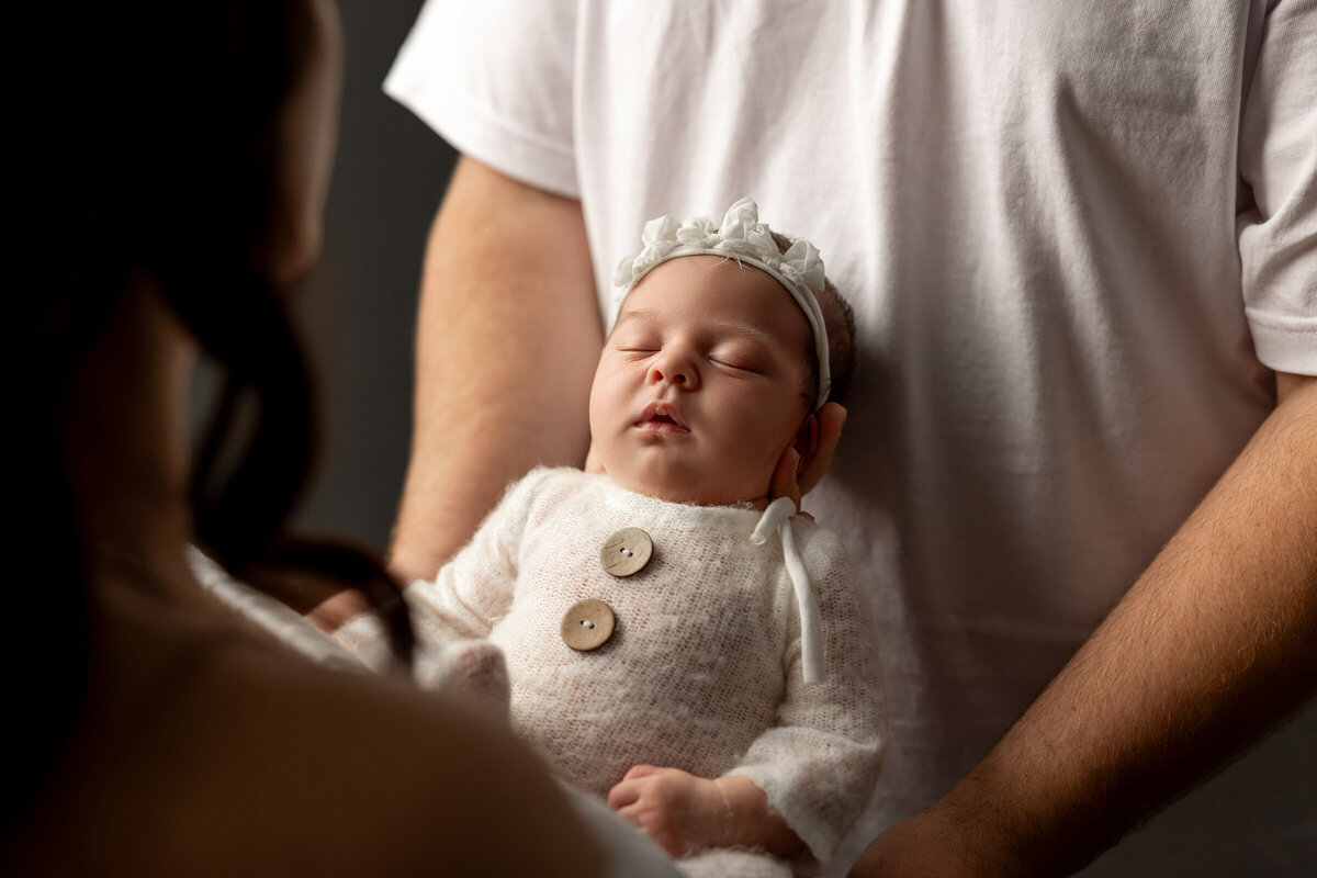 Newborn baby girl swaddled in a cozy white knit outfit with button details and a matching headband, cradled gently in her parent's arms. Captured by Inland Empire newborn photographer Kaitlyn Dawn Photography.
