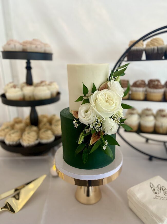 A two tier cake with a white tier on top and an emerald green tier on the bottom. It's decorated with a cluster of white flowers and fresh figs on the side between the two tiers. In the background is two stands full of cupcakes slightly blurred.