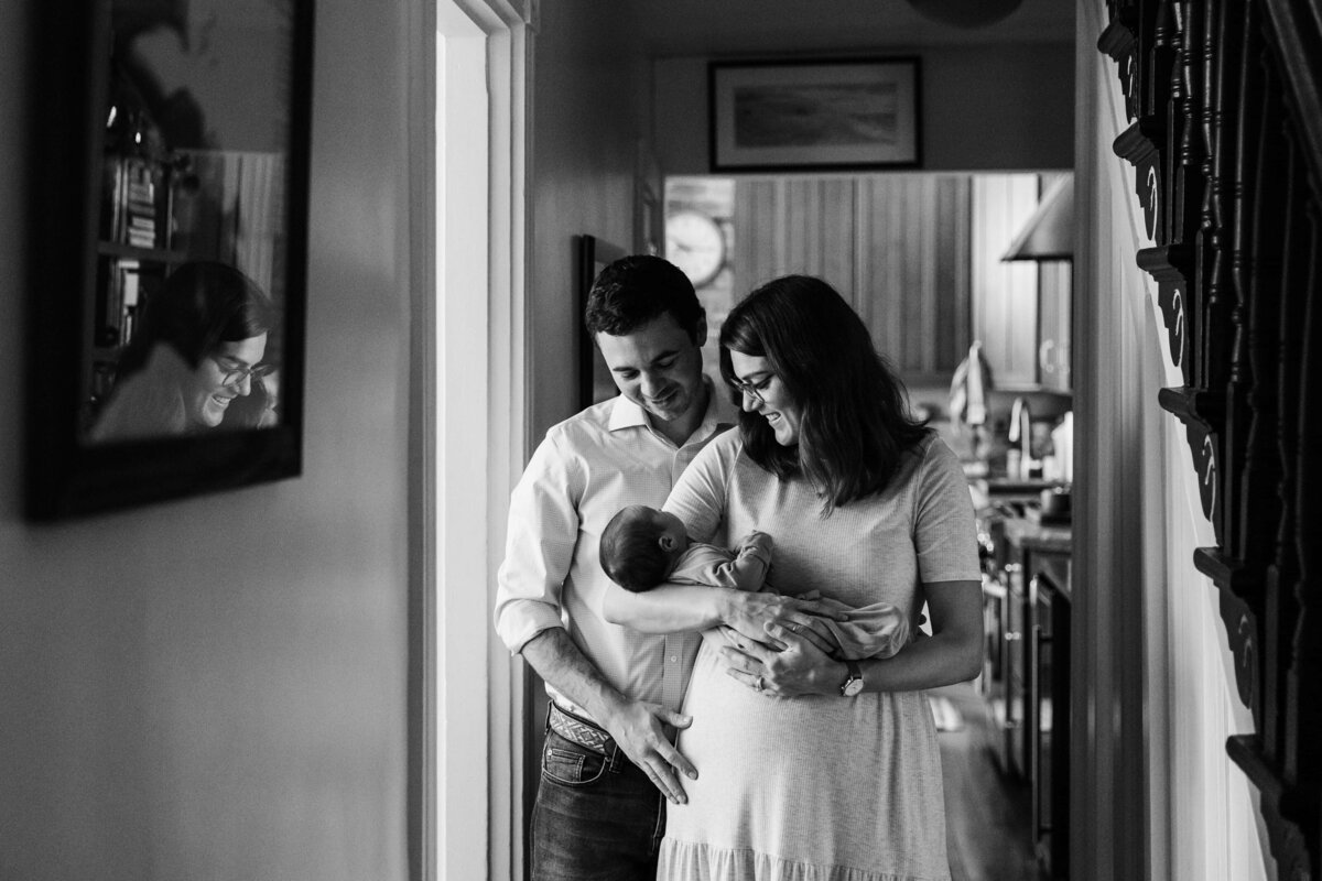 A black and white photo of a smiling couple holding a baby at home, captured during a home newborn photography session.