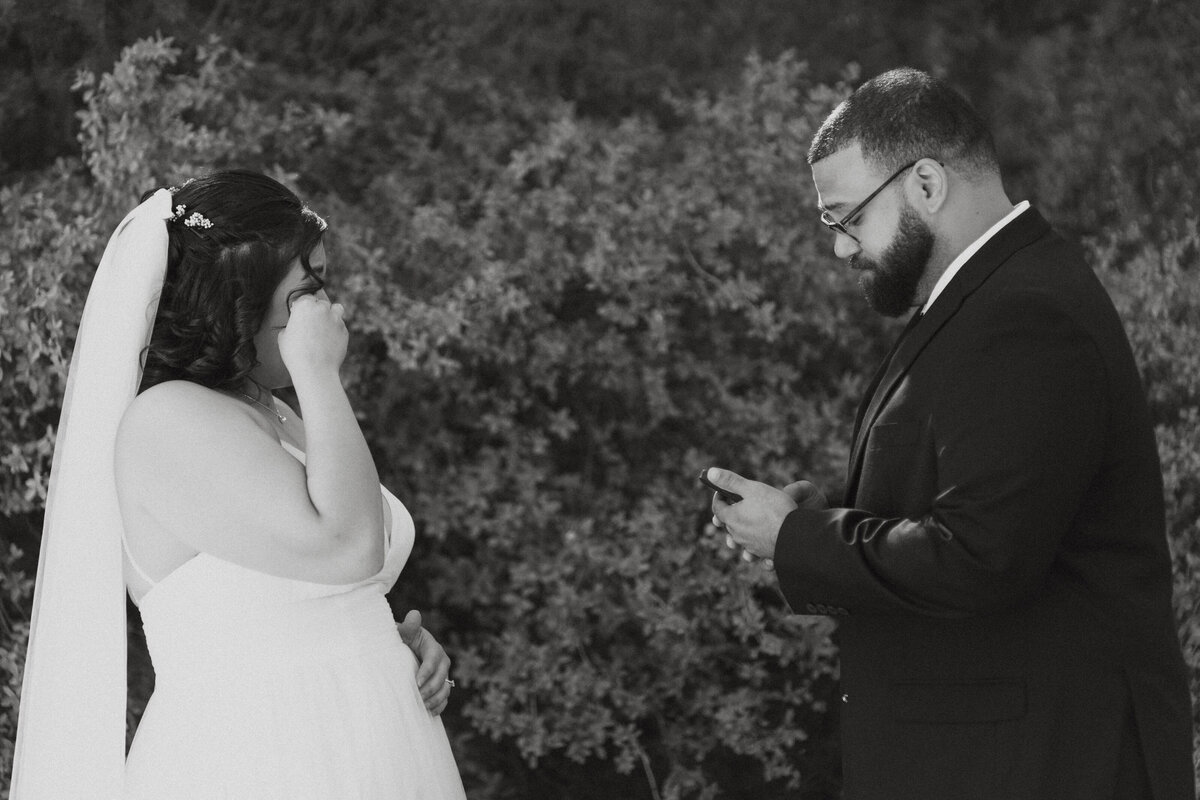 A newlywed couple standing together as one reads a note to the other 
