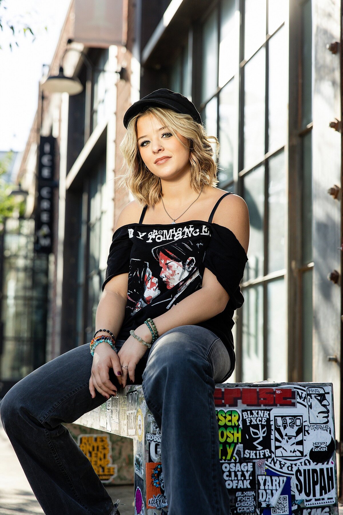 Urban senior portrait with edgy vibes, teen girl in graphic tee and jeans sitting on a sticker-covered bench in Los Angeles’ Arts District
