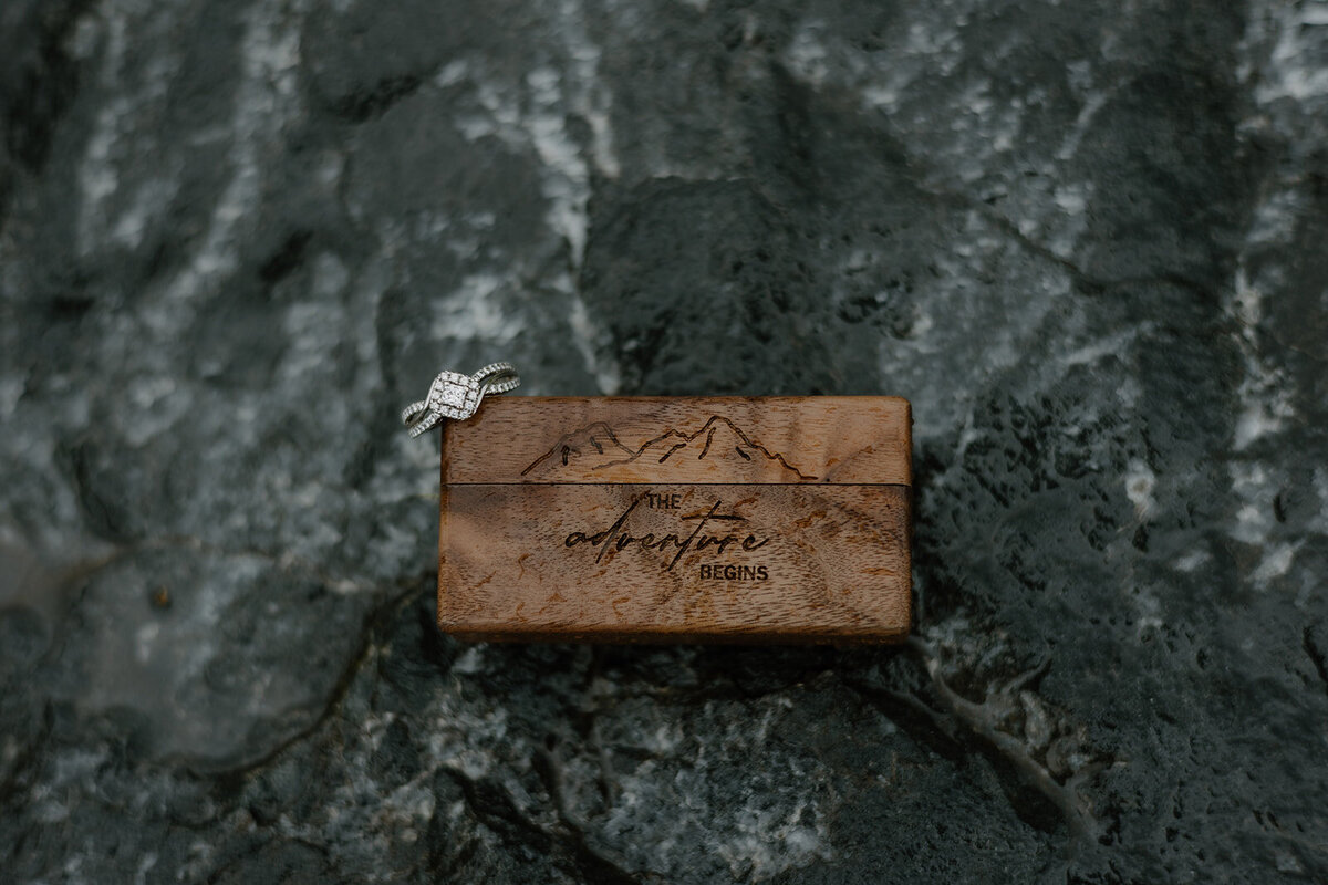 Engagement ring balancing on the edge of wooden ring box, placed on rock.