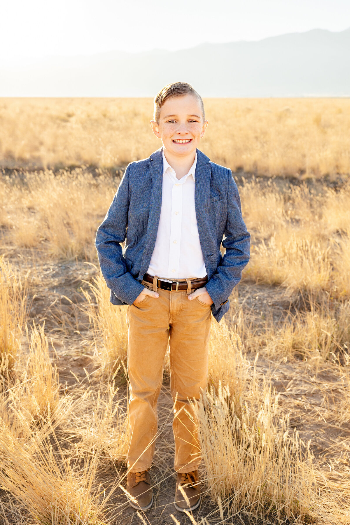 A young boy wearing a blue blazer and tan pants smiles confidently while standing in a golden field in Eagle Mountain, Utah, during a sunset family photoshoot.