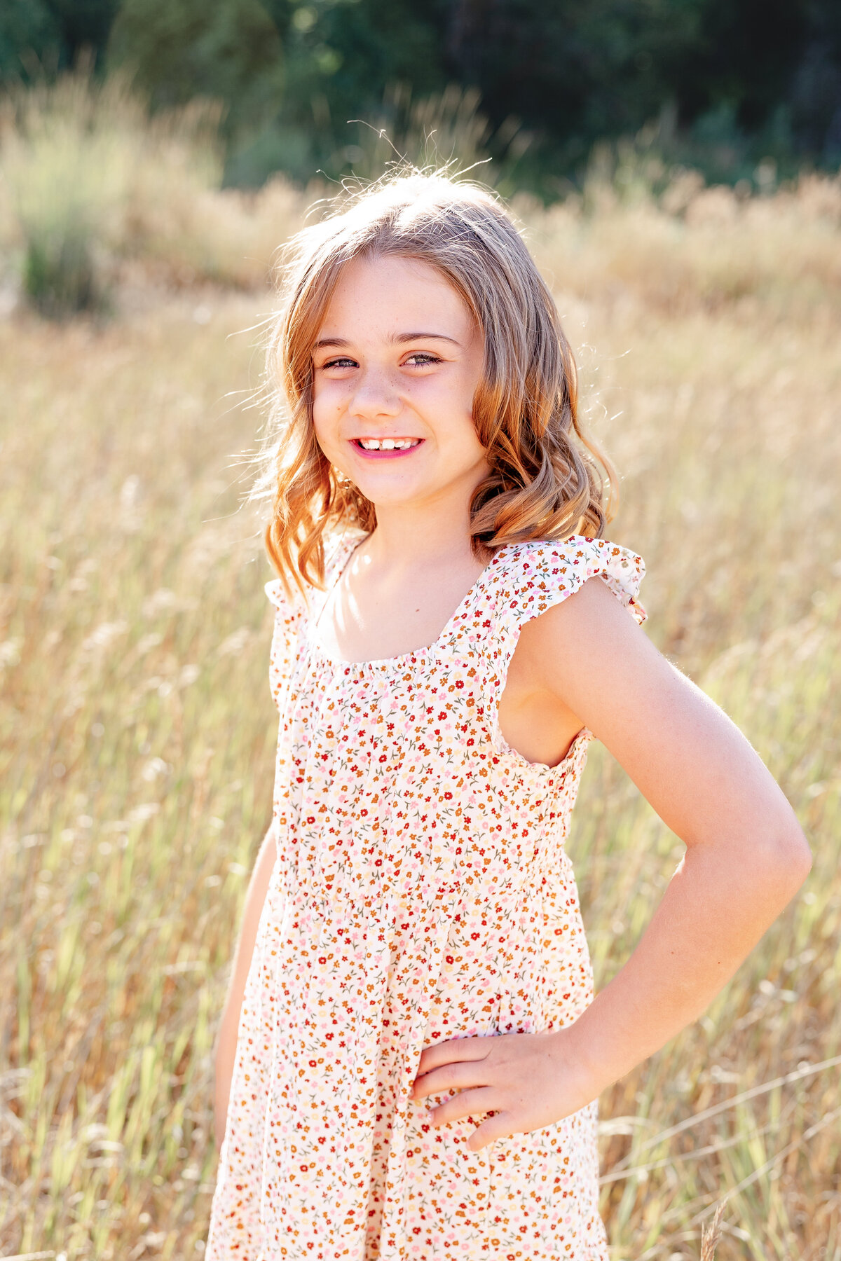 A girl with light brown hair poses in a sunlit meadow, wearing a floral dress with delicate ruffled sleeves, radiating warmth and happiness.