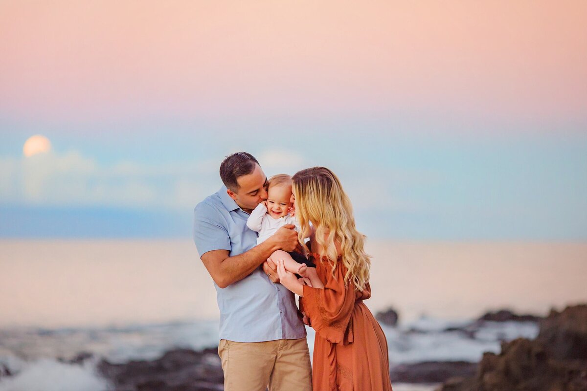 Woman in burnt umber dress and blond wavy hair leans in to kiss her young child on Maui under the full moon