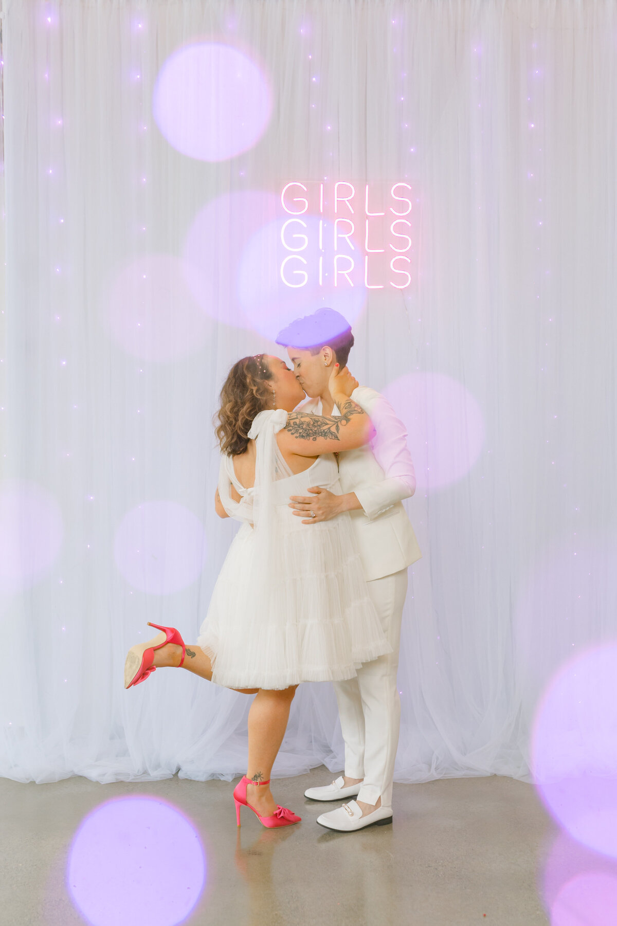 Two brides kissing with a white backdrop and a "Girls, girls. girls" neon sign in the background