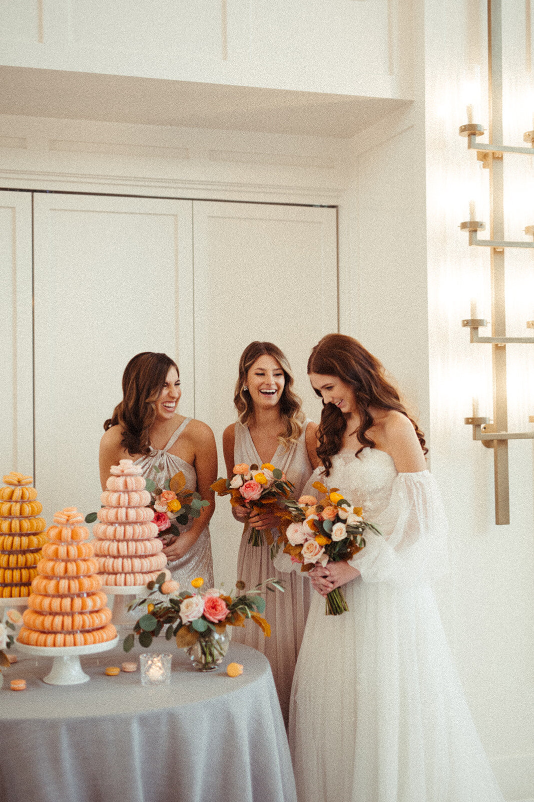 A bride wearing a white wedding gown holds a bouquet of flowers with two women smiling next to a table filled with macaroons.