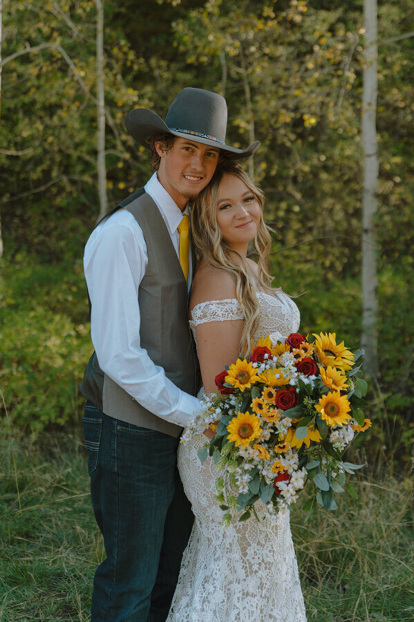 A groom holds his bride from behind, both smiling at the camera for a portrait. She's holding her wedding bouquet full of sunflowers.