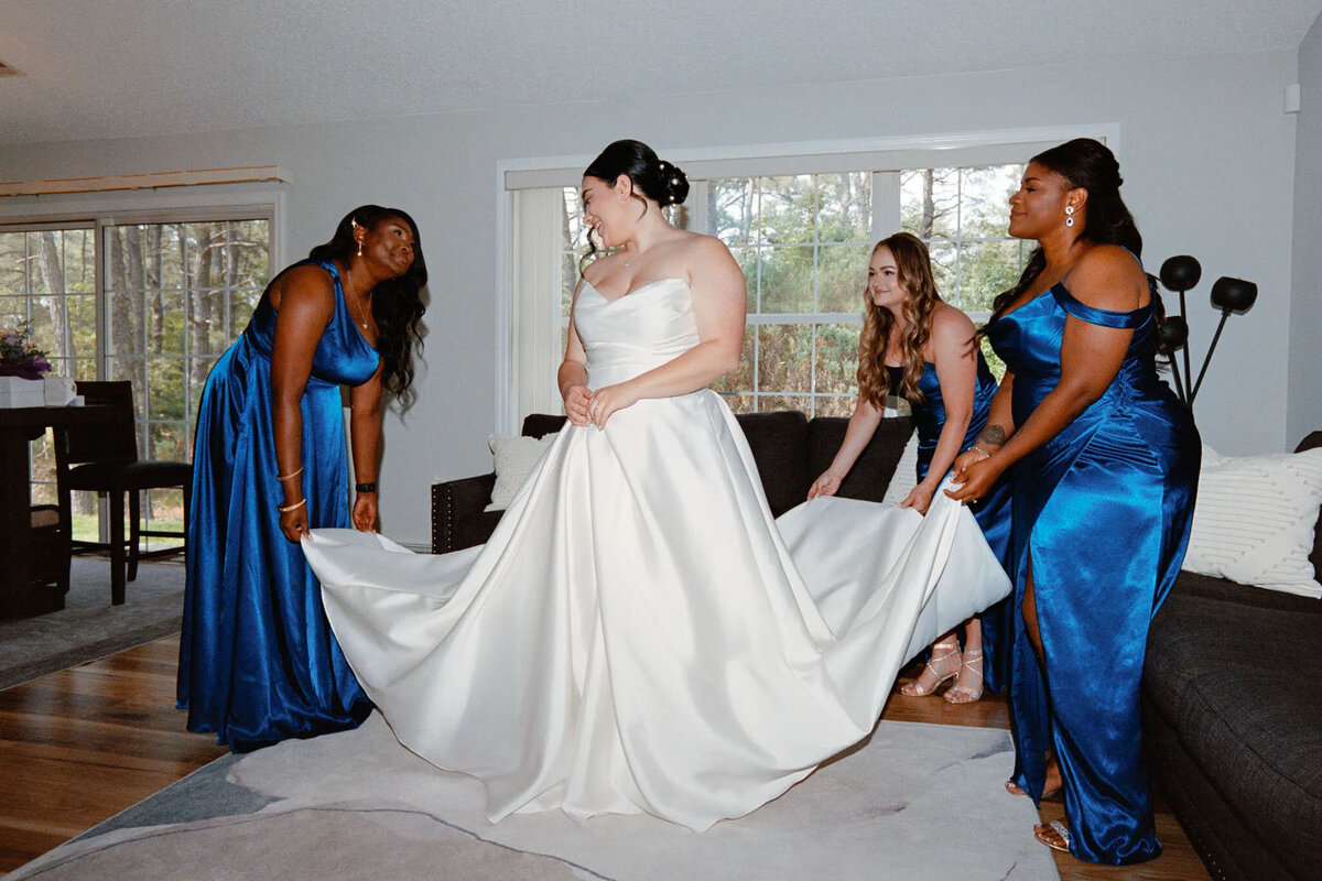 A bride in a white gown stands smiling as three bridesmaids in blue dresses help arrange her dress in a bright living room, perfectly captured by an attentive NJ wedding photographer.
