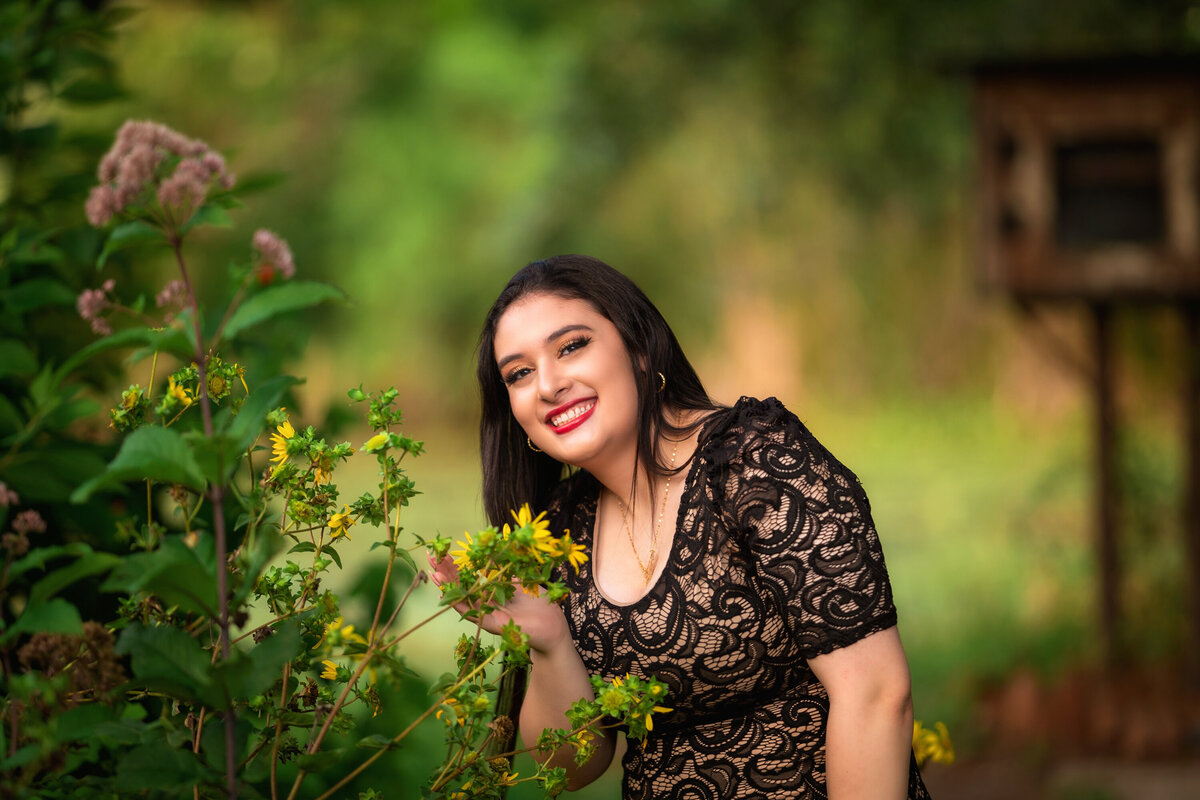 High school senior girl in a black lace dress.  She is middle eastern with long, dark black hair.  She is smiling and wearing red lipstick.  She is near a bush of flowers and is reaching to smell a yellow flower.  There are blurry trees behind her.