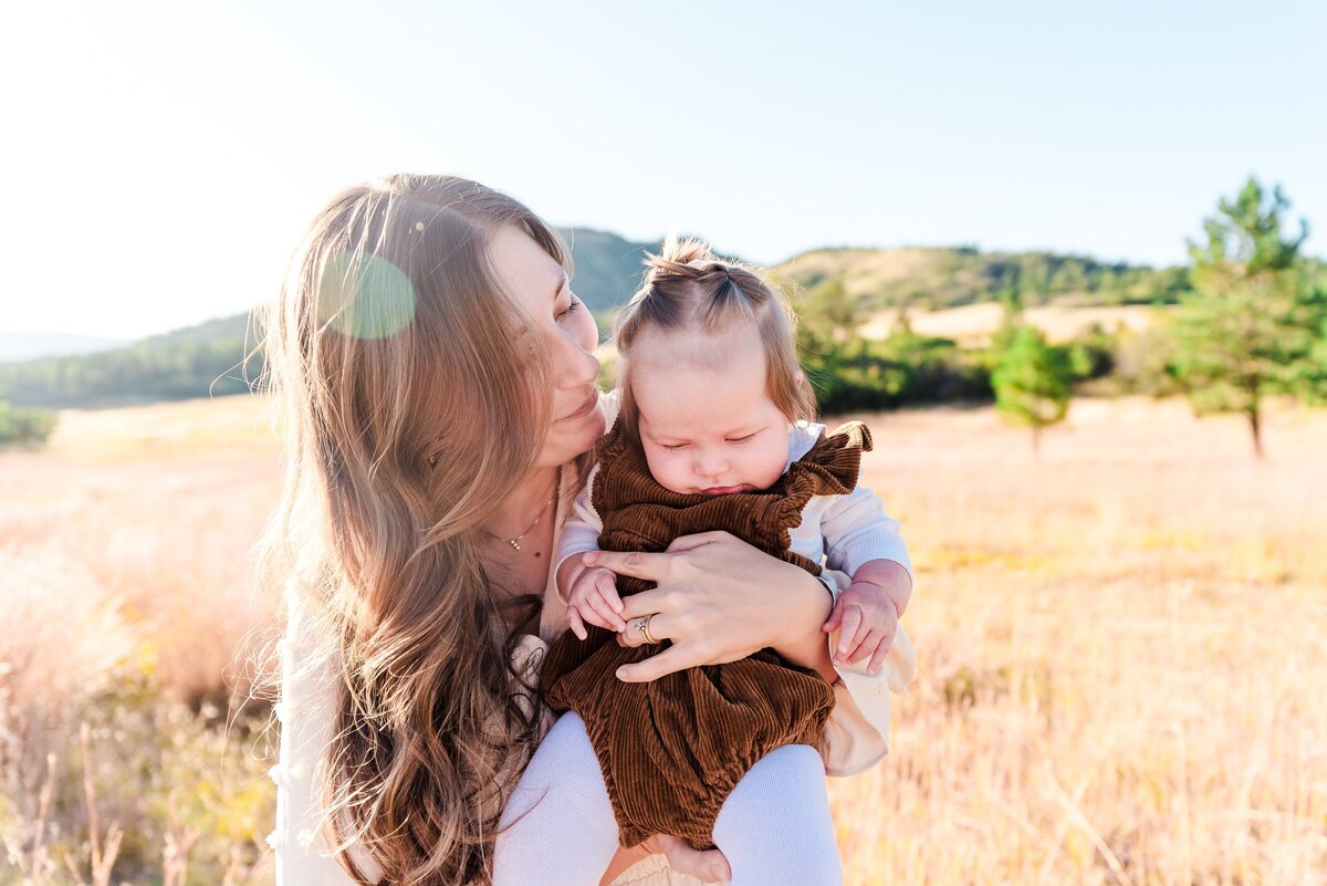 a mom with long brown hair holds her infant daughter in her arms as she leans in toward her daughter and a sun glare goes through her hair captured by denver family photographer