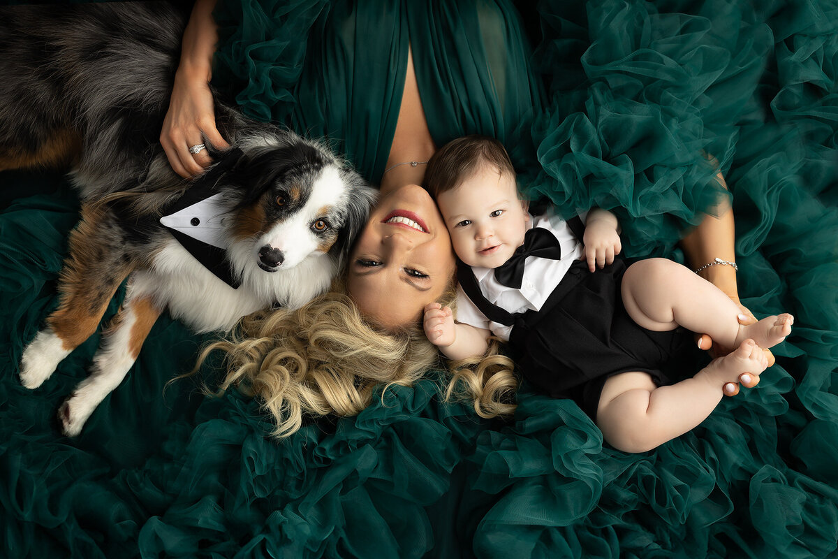 Dallas milestone photographer captures a radiant mother lying on emerald green tulle with her smiling baby in a tuxedo romper and their dapper dog, all dressed to impress.