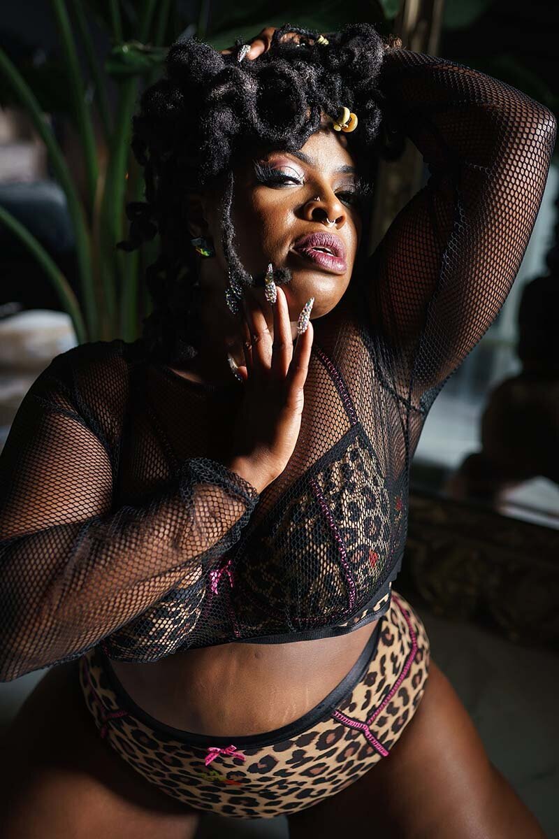Boudoir portrait of dark skinned woman kneeling wearing a leopard lingerie set and fishnet top kneeling with her hand on her face
