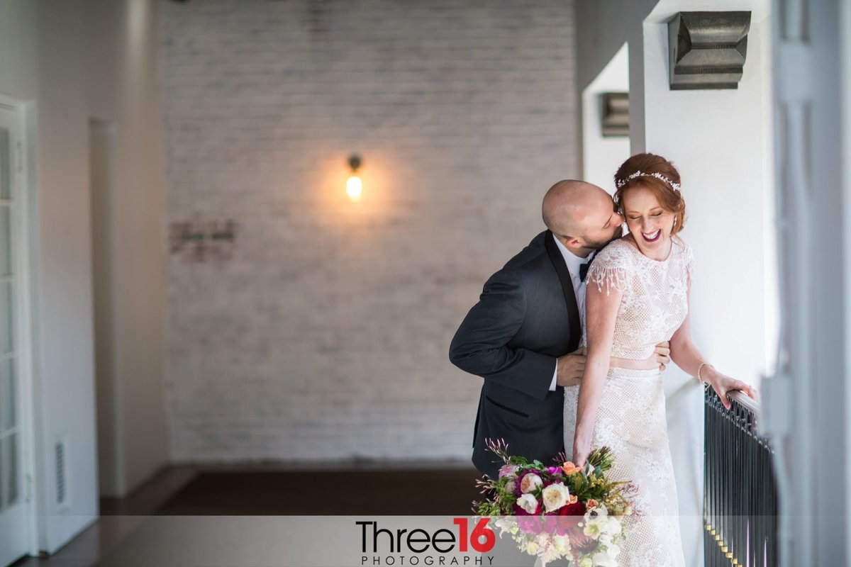 Groom hold his Bride from behind and whispers in her ear causing her to laugh