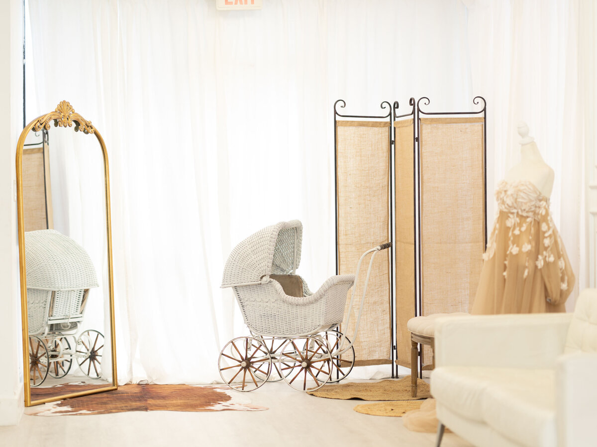Vintage room with a white wicker baby carriage, ornate mirror, and beige dress on a mannequin. Background includes a folding screen and white couch.