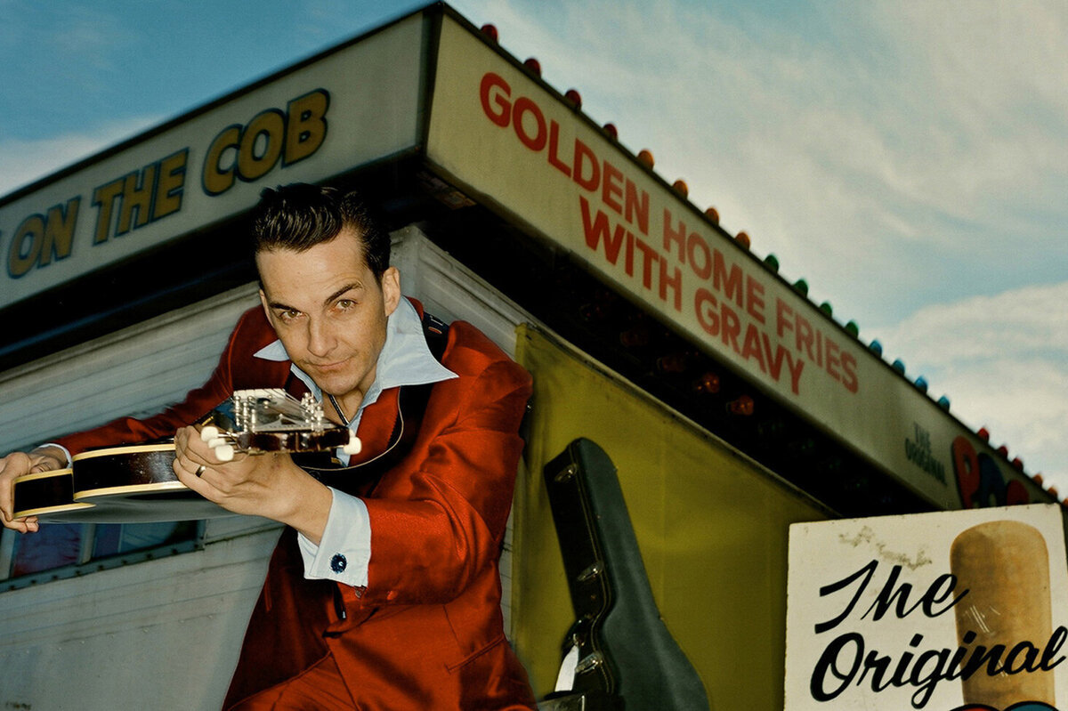Musician portrait Rich Hope wearing red suit with blue shirt holding guitar toward camera in front of food stand wooden folding sign beside him Just Play Something Gallery