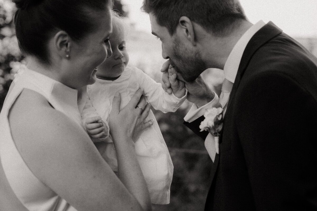 Un instant tendre entre le marié et son enfant. Un doux baiser sur la main sous le regard attendri de la mariée. Photographie de mariage par Kamescop.