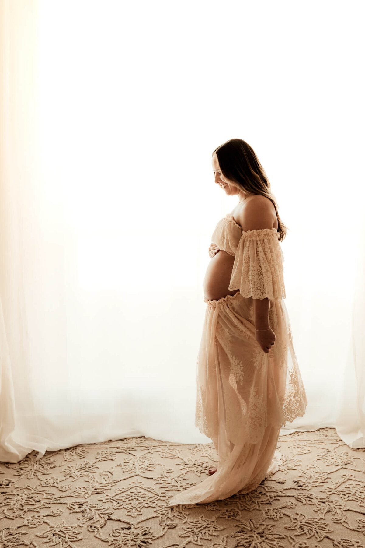 Expecting mother smiles down at her baby bump while standing near a window and wearing a blush pink lace dress.